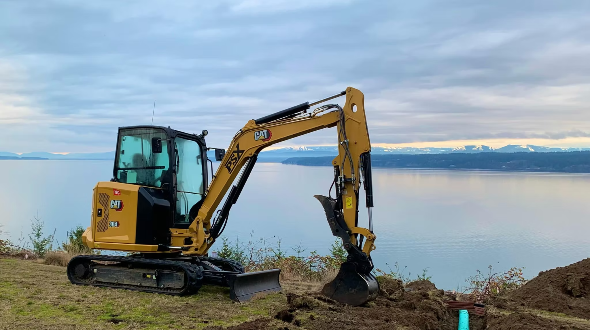 A yellow CAT 304 mini excavator digging soil near a body of water with mountains visible in the background
